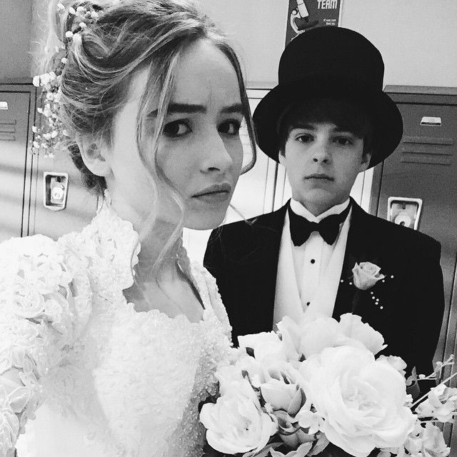 Peyton Meyer in a black formal dress with a black hat and Sabrina Carpenter in a white bridal gown holding white flowers.